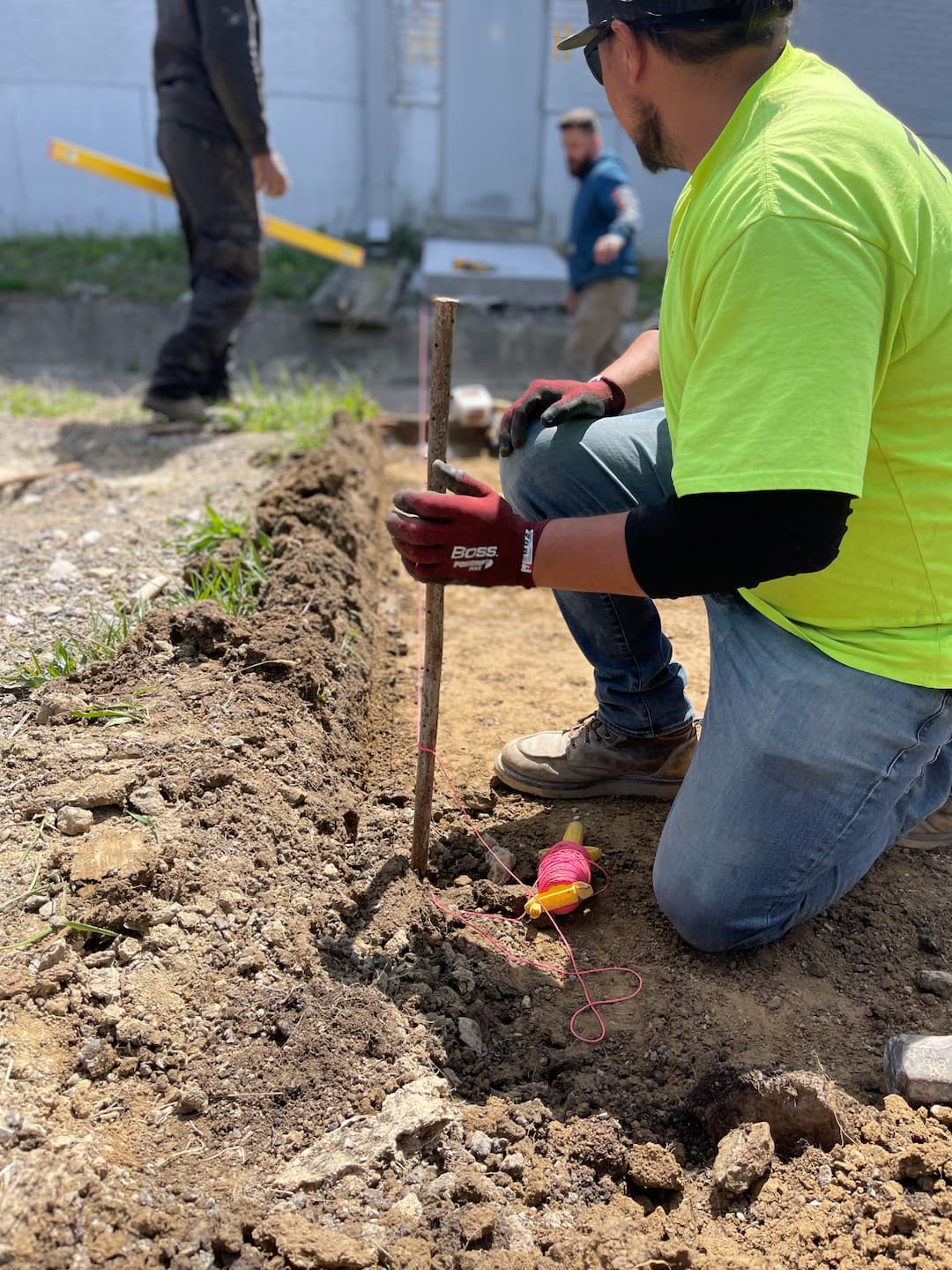Wesney facility maintenance crew member kneeling in a freshly excavated trench, setting a wooden grade stake with mason's twine to establish line and elevation for site work.
